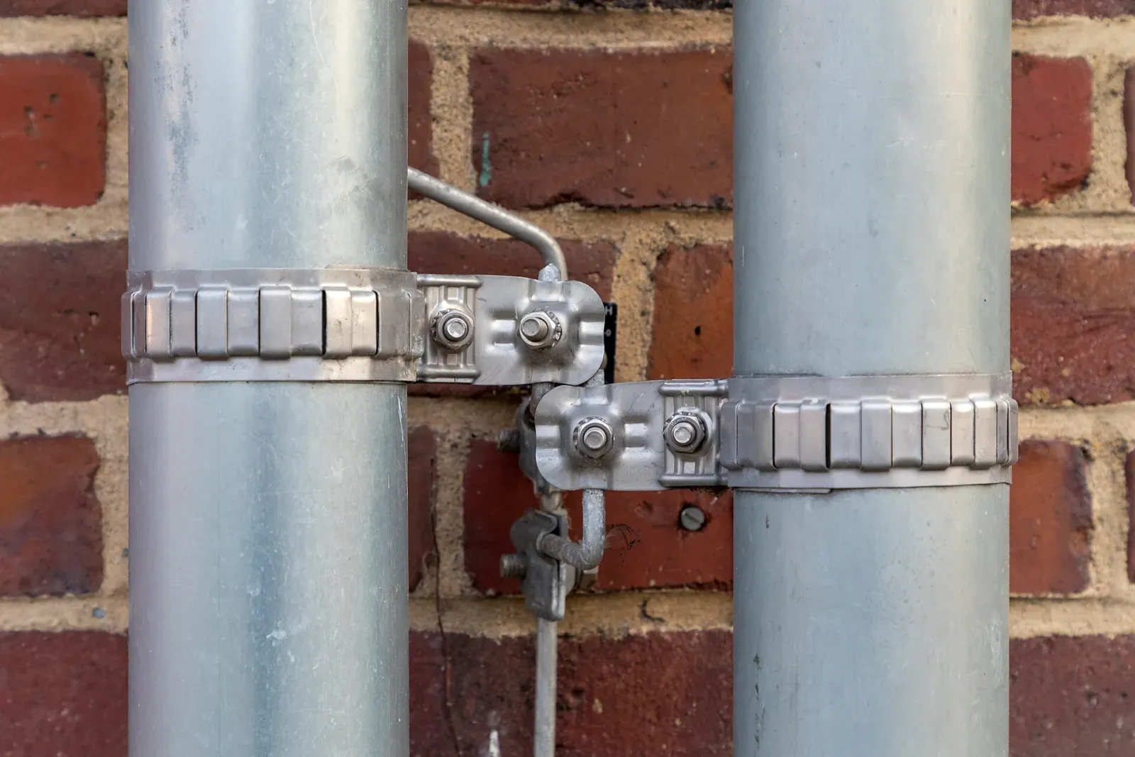 Stainless bonding clamps joining two metal downspouts to a lightning protection conductor on a brick wall