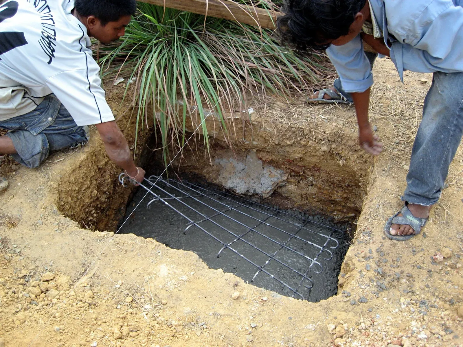 Two workers lowering a rebar mesh into a footing excavation filled with wet concrete