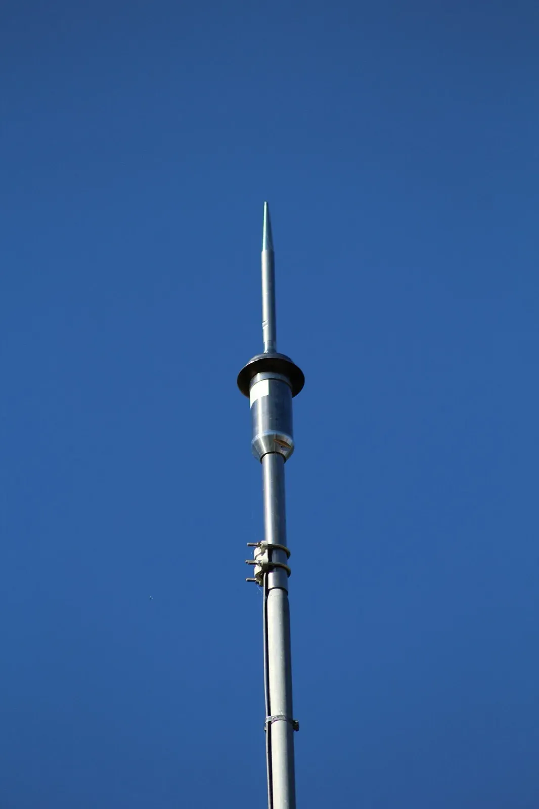 Single lightning rod on a tall metal mast with a cylindrical head below the tip, against a clear blue sky