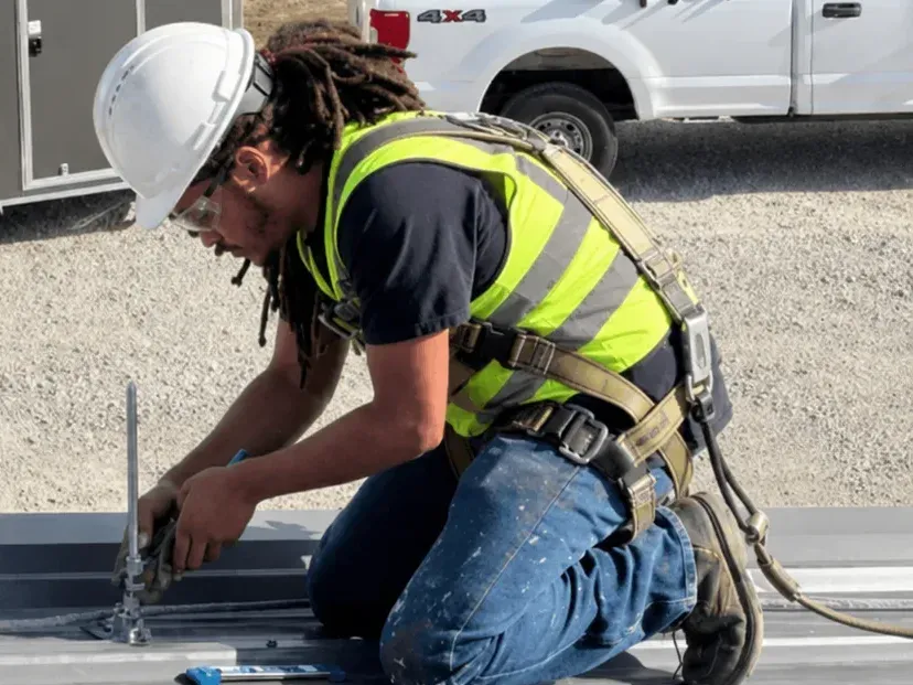 Site Safe Lightning Protection technician in a hard hat and harness fastening an air terminal base on a metal roof