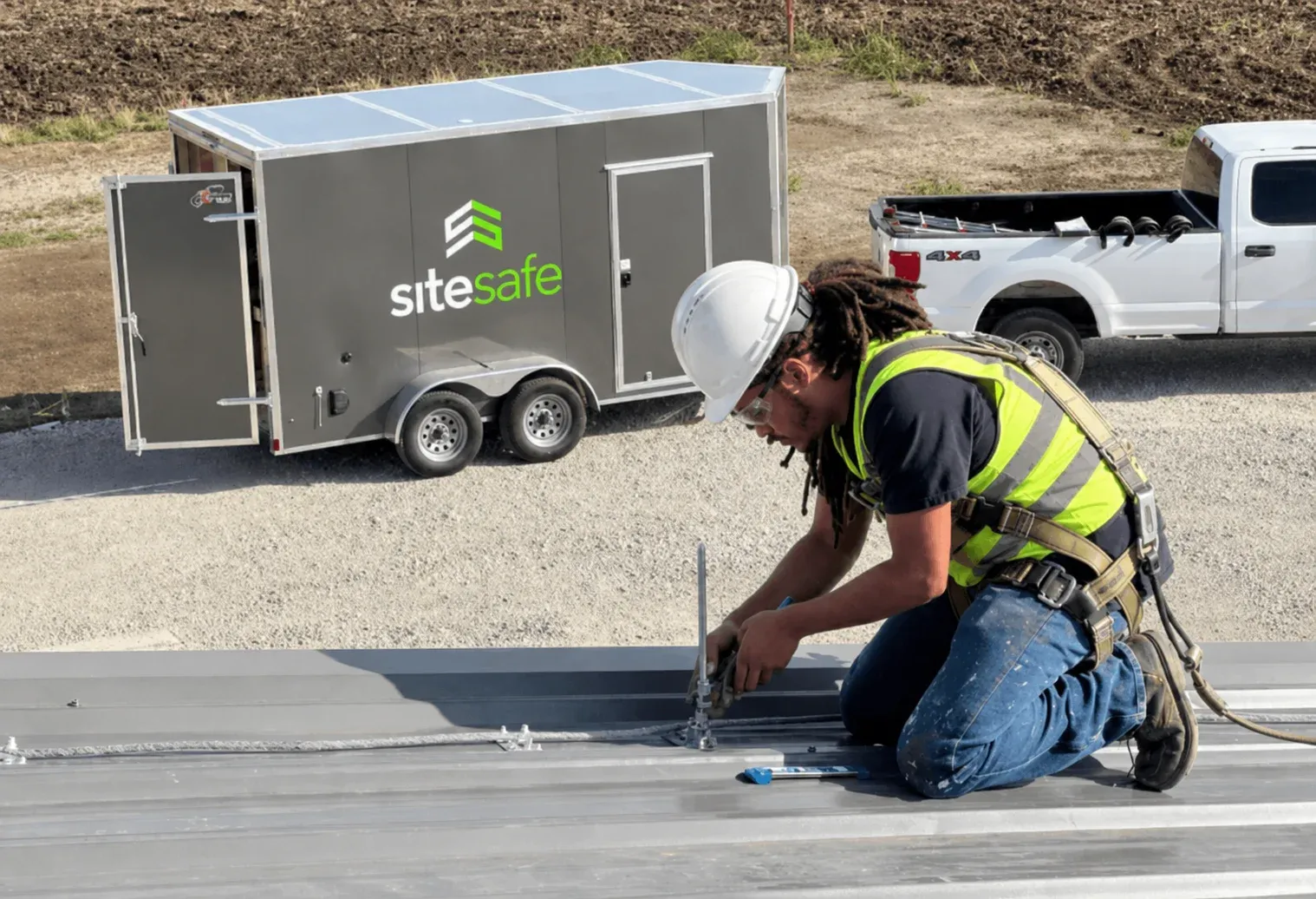 Technician in a hard hat and harness installing an air terminal on a metal roof, with a work trailer and pickup truck below